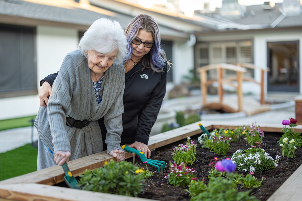 PACS-300x200-activity2 auburn oaks female staff member assisting an elderly woman while she digs with a shovel in a raised flower bed.