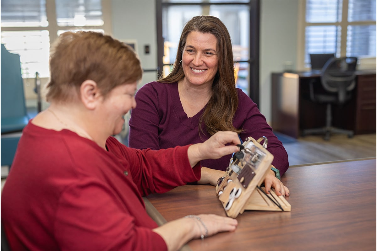 PACS-300x200-caregiver2 Auburn oaks female staff member smiling at woman as she uses a sensory and motor skills board.